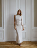 A woman wearing a sleeveless, off-white midi dress with draped shoulder details and a side cutout, standing in front of a white paneled door on a wooden floor.